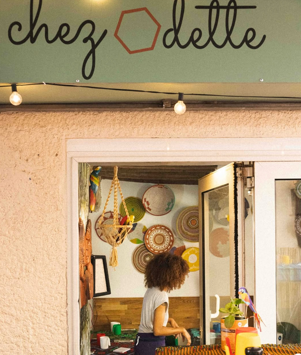 Une femme travaille à l'intérieur d'un café confortable au décor coloré, dont la vitrine porte l'inscription "chez dette". Limbus Studio