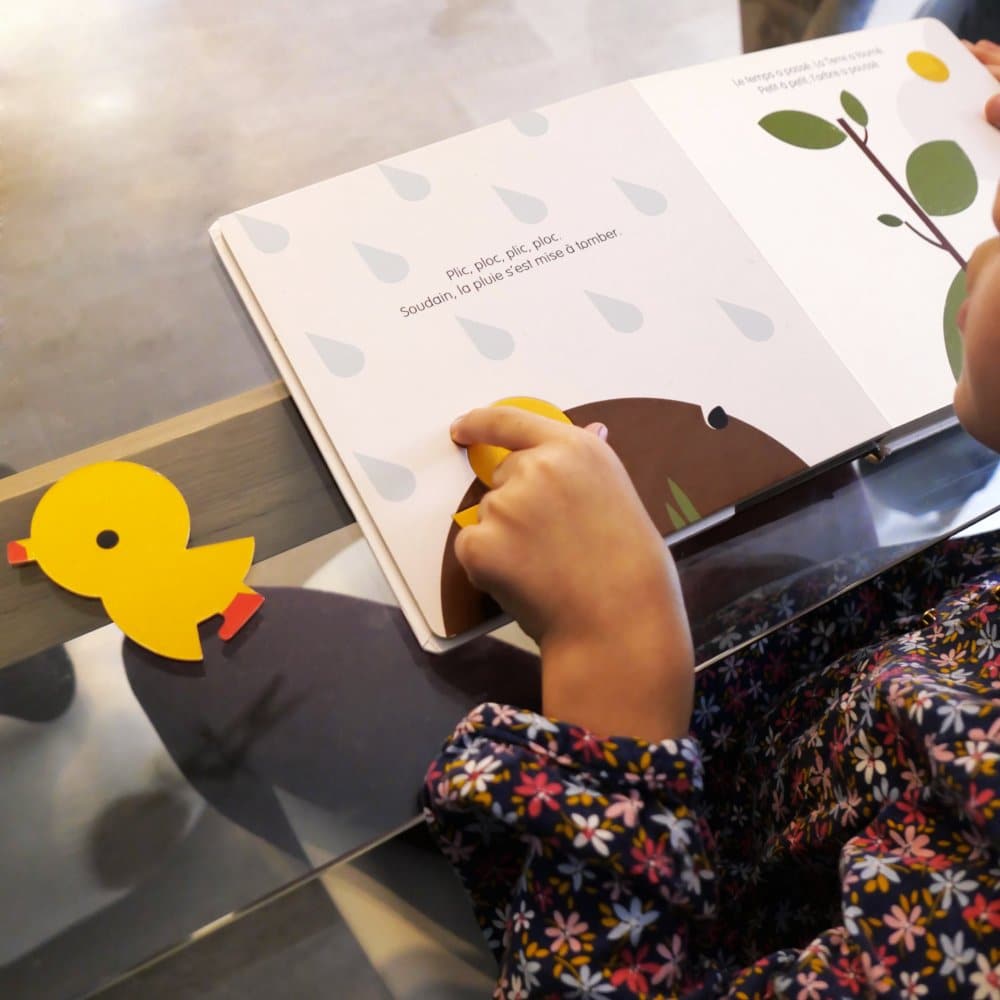 Un enfant plongé dans un livre d'images est assis à une table, où repose un charmant canard en papier, donnant vie au monde de l'imagination. Dans cette scène chaleureuse, la créativité coule librement comme si elle était guidée par la main d'un artisan dans une agence de création. Limbus Studio