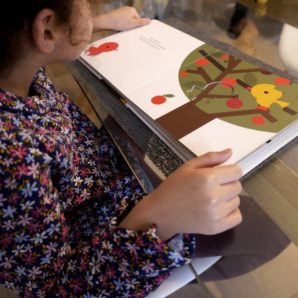 Dans un studio de design chaleureux, un enfant est assis à une table, plongé dans un livre d'images sur les pommes. Limbus Studio