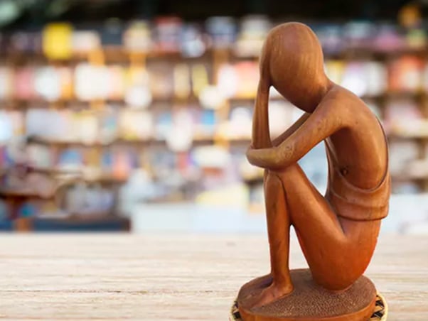 Sculpture en bois d'une figure pensive assise, avec un arrière-plan de bibliothèque flou, parfaite pour rehausser le design de votre studio. Limbus Studio