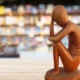 Sculpture en bois d'une figure pensive assise, avec un arrière-plan de bibliothèque flou, parfaite pour rehausser le design de votre studio. Limbus Studio