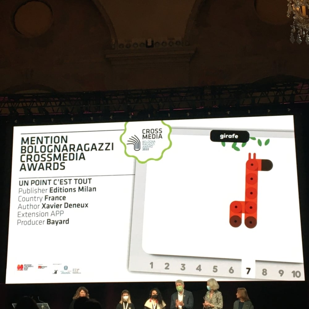 Un groupe sur scène présente un prix aux BolognaRagazzi CrossMedia Awards, sous une illustration de girafe saisissante qui met en valeur une créativité et un design exceptionnels. Limbus Studio