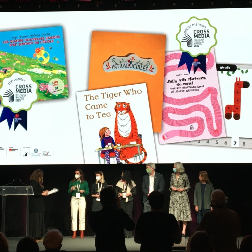 Des gens sur scène lors d'une cérémonie de remise de prix avec des livres affichés sur un écran au-dessus d'eux, mettant en valeur le summum de la création littéraire et du design innovant. Limbus Studio