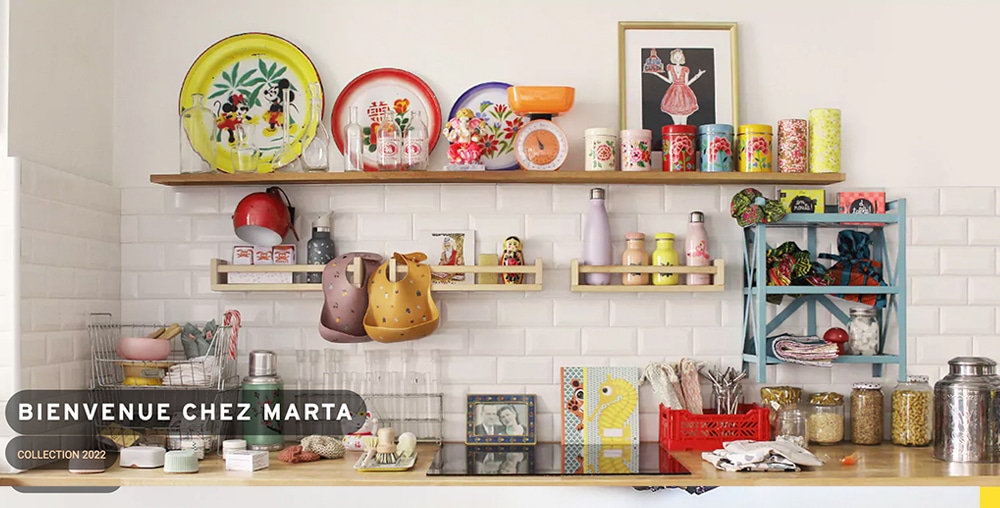 Étagères de cuisine colorées avec plats, tasses et objets décoratifs, arborant un panneau « Bienvenue chez Marta » - un design de studio ravissant qui illumine n'importe quel espace. Limbus Studio
