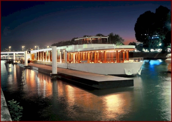 Un bateau fluvial moderne aux larges fenêtres, symbole d'un design élégant, est amarré la nuit. Les doux reflets de ses lumières dansent sur l'eau calme, révélant une création magistrale de l'agence à l'origine de ce navire époustouflant. Limbus Studio