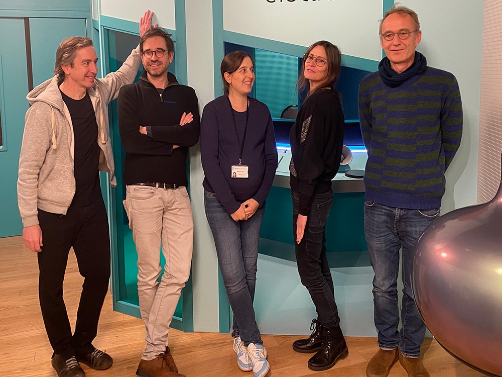 Cinq personnes debout dans une pose décontractée, souriantes, contre un mur bleu sarcelle à l'intérieur, donnant vie à une ambiance de studio dynamique. Limbus Studio
