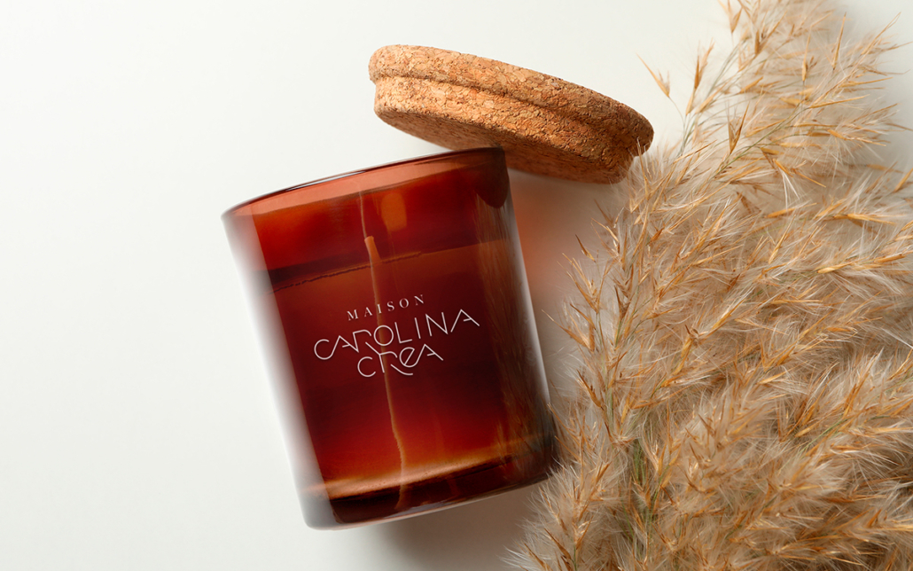 La bougie marron de Maison Carolina dans un pot en verre avec un couvercle en liège se trouve élégamment à côté de l'herbe de la pampa séchée sur une surface blanche, incarnant le style signature de la marque en matière de sophistication discrète. Limbus Studio