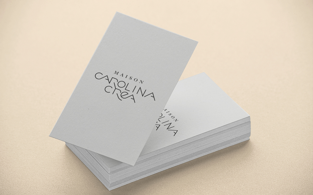 Une pile de cartes de visite sur fond beige affiche le texte « MAISON CAROLINA CREA » dans une police élégante et moderne.