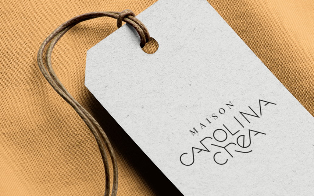 Un tissu beige avec une étiquette beige-gris étiquetée « Maison Carolina Crea » attachée par une ficelle, présentant un design exquis et une mode intemporelle. Limbus Studio