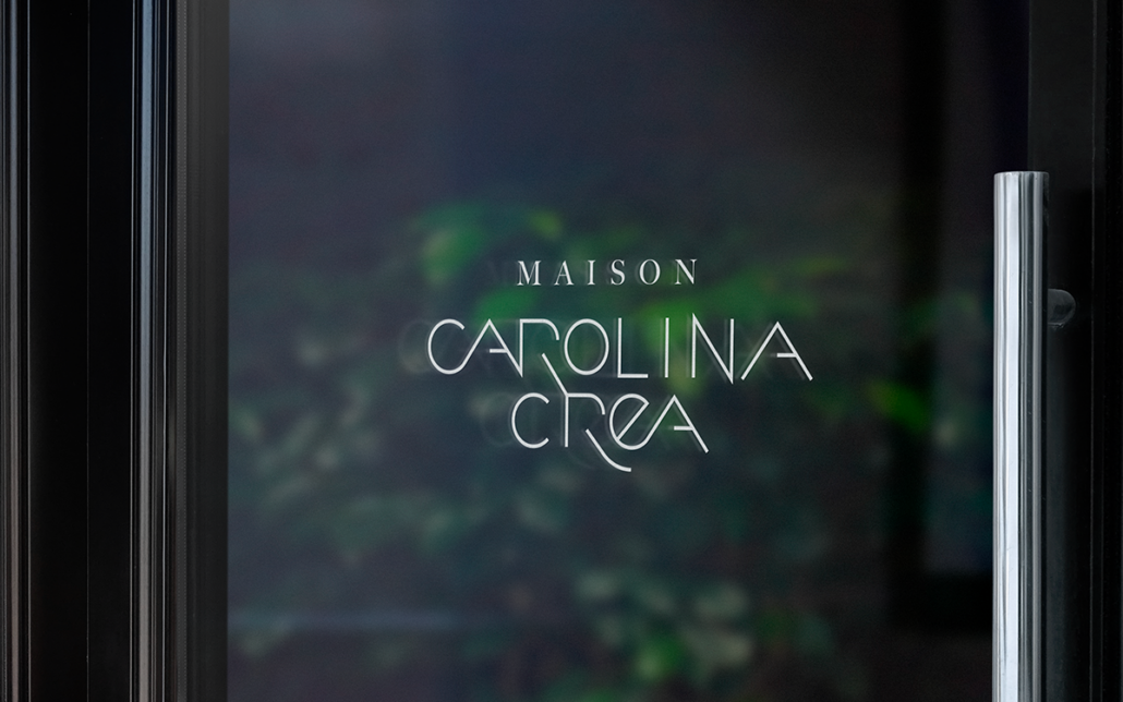 Une porte vitrée affichant le texte « Maison Carolina Crea », reflétant une verdure luxuriante, laisse entrevoir une agence de design innovante où les idées créatives fleurissent comme la nature. Limbus Studio