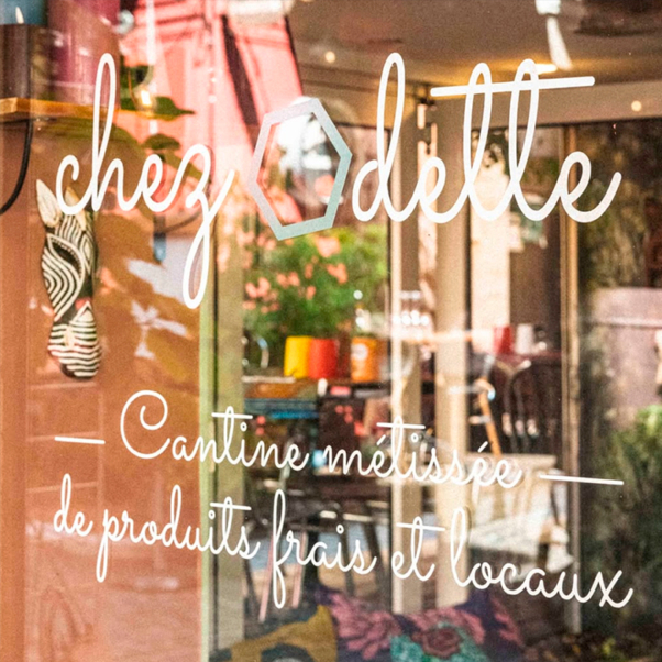 Porte en verre avec écriture blanche indiquant "chez Odette, Cantine métissée de produits frais et locaux", reflétant l'intérieur d'un café. Pour en savoir plus sur chez odette, consultez le site chezodette.com. Limbus Studio