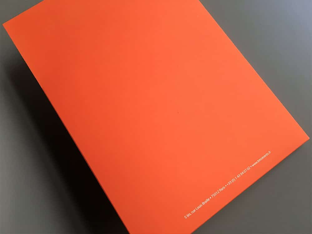 Un livre orange repose sur une surface grise, son design élégant complété par un petit texte blanc dans le coin inférieur. Limbus Studio
