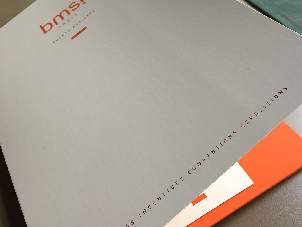 Dossier gris aux accents orange, marqué « bms », présentant un design élégant et un texte sur les réunions et les expositions sur le côté. Limbus Studio