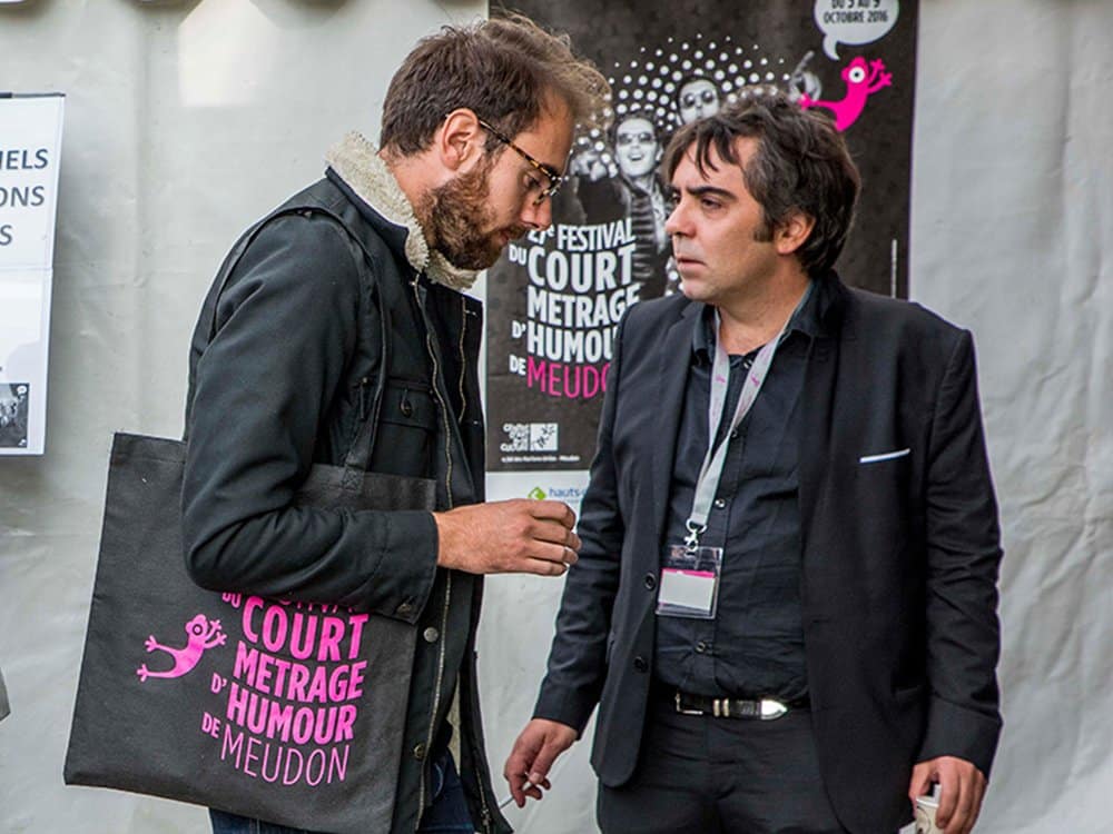 Deux hommes conversent lors d'un festival de courts métrages comiques, l'un tenant un sac fourre-tout à thème conçu avec un graphisme ludique, des affiches en arrière-plan présentant une création vibrante. Limbus Studio