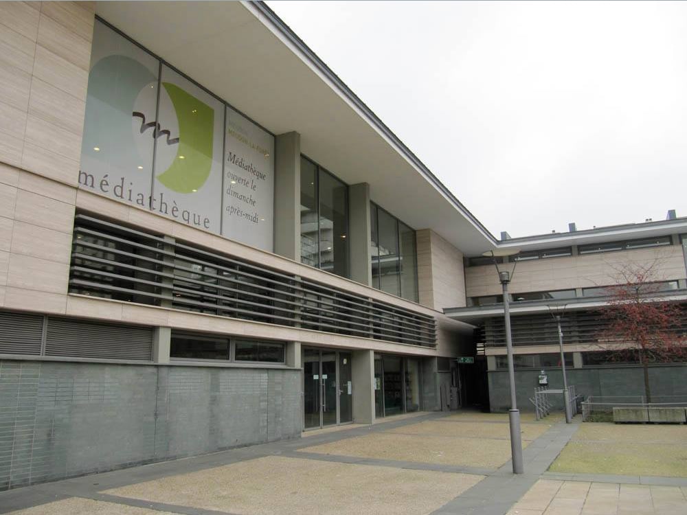 Le bâtiment moderne de la bibliothèque, avec son design élégant et ses grandes baies vitrées, affiche fièrement un panneau indiquant « médiathèque », invitant les visiteurs à entrer. Limbus Studio