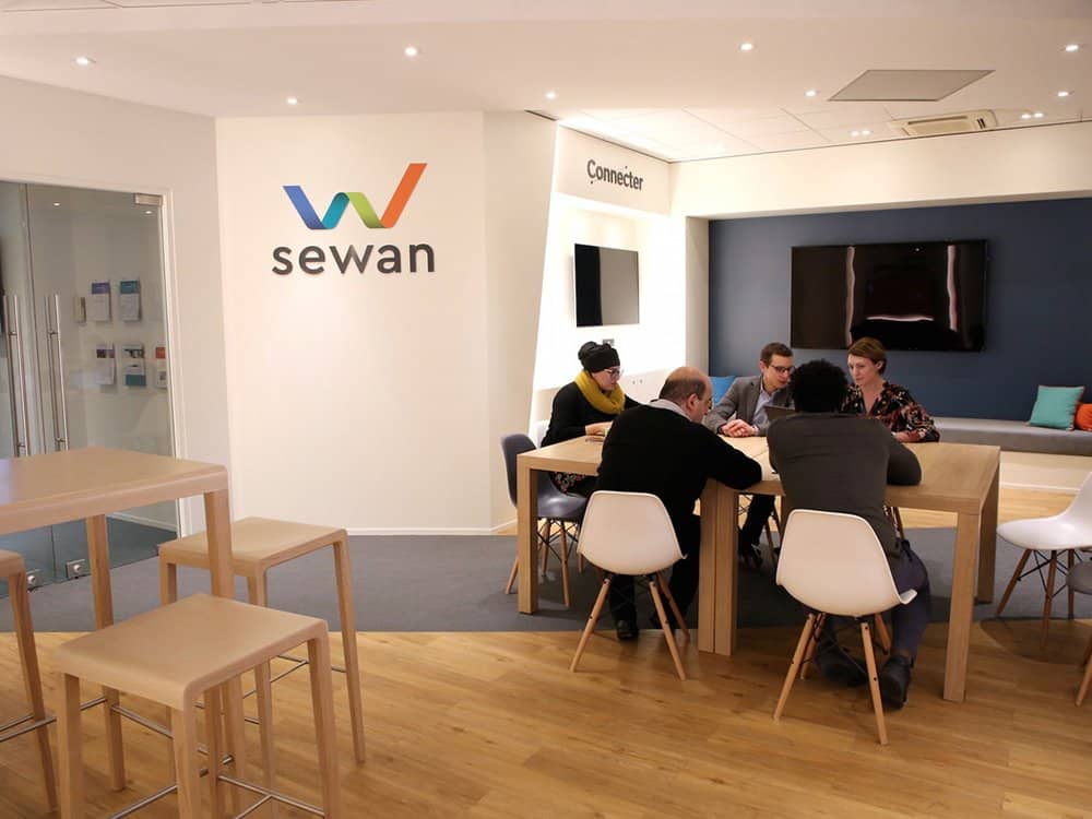 Des personnes assises à une table dans un bureau de studio moderne avec le logo « sewan » sur le mur, discutant de leur dernière création sur WordPress. Limbus Studio