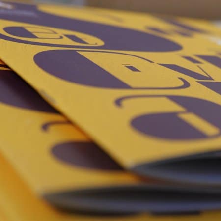 Gros plan de brochures empilées présentant un design accrocheur avec un grand texte violet audacieux sur un fond jaune vif. Limbus Studio